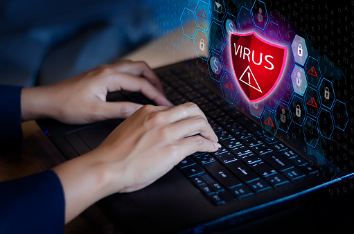 A person typing on a laptop keyboard. The laptop screen has a large red image on it that says Virus