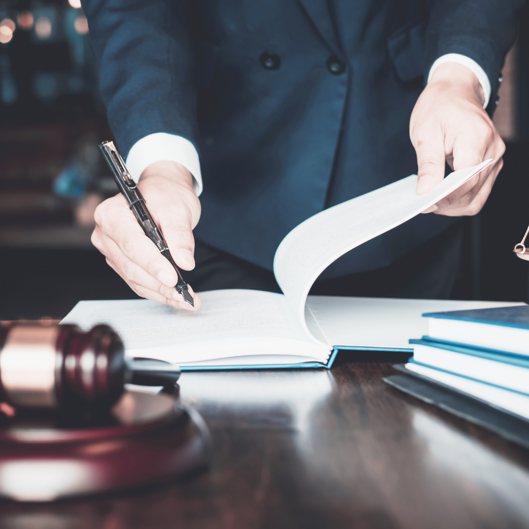 A lawyer, dressed in a formal suit, is seen reviewing and marking documents at a desk. The scene includes a wooden gavel and several open books, symbolizing legal work and decision-making. The lawyer’s hands are focused on the paperwork, highlighting the meticulous nature of legal practices.